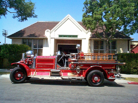 Vintage red fire engine with chrome fittings and wooden ladder rack parked in front of a white firehouse
