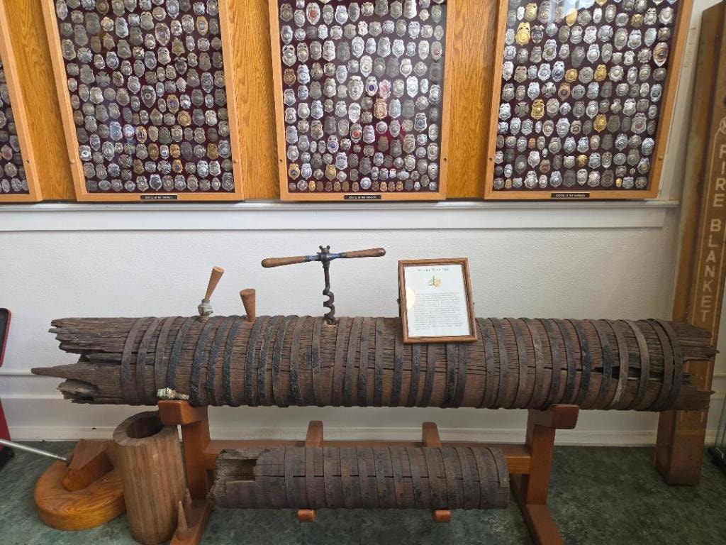 Historic wooden water pipe on a display stand with iron banding, with a large badge collection donated by Ray Chambers mounted on the wall behind it